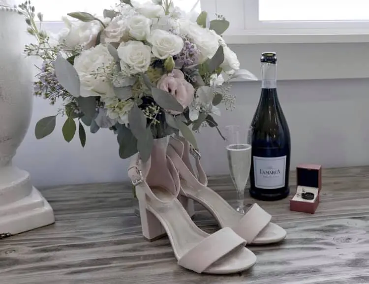 white floral bouquet is displayed along with bridal shoes, a glass and bottle of champagne, and the wedding ring