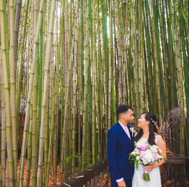 bride holding floral bouquet faces her groom against tall wall of bamboo